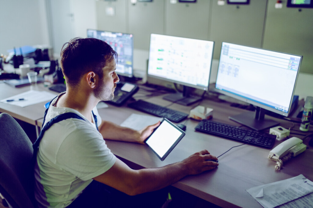 Hardworking plant worker sitting in control room, holding tablet and using computer for monitoring.